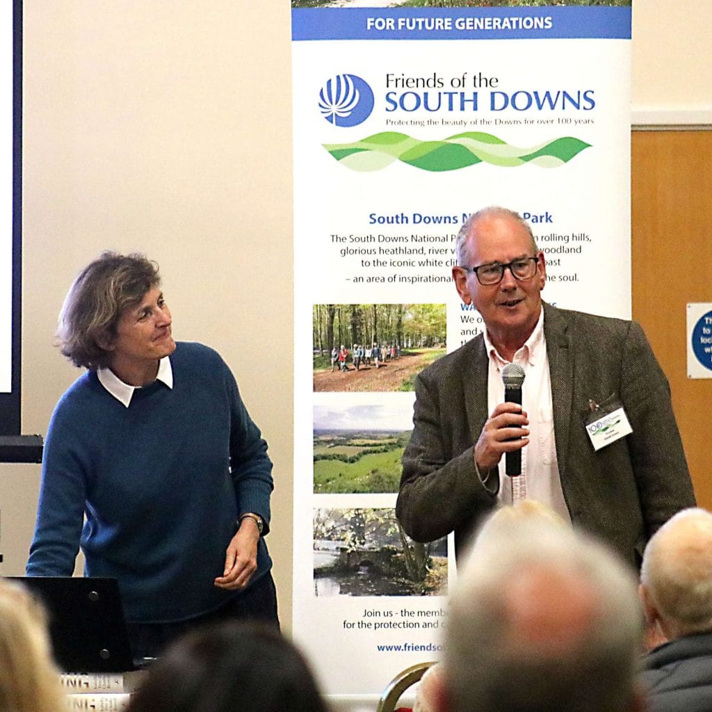 A man and woman address a crowd, standing together in front of an audience at a public gathering.