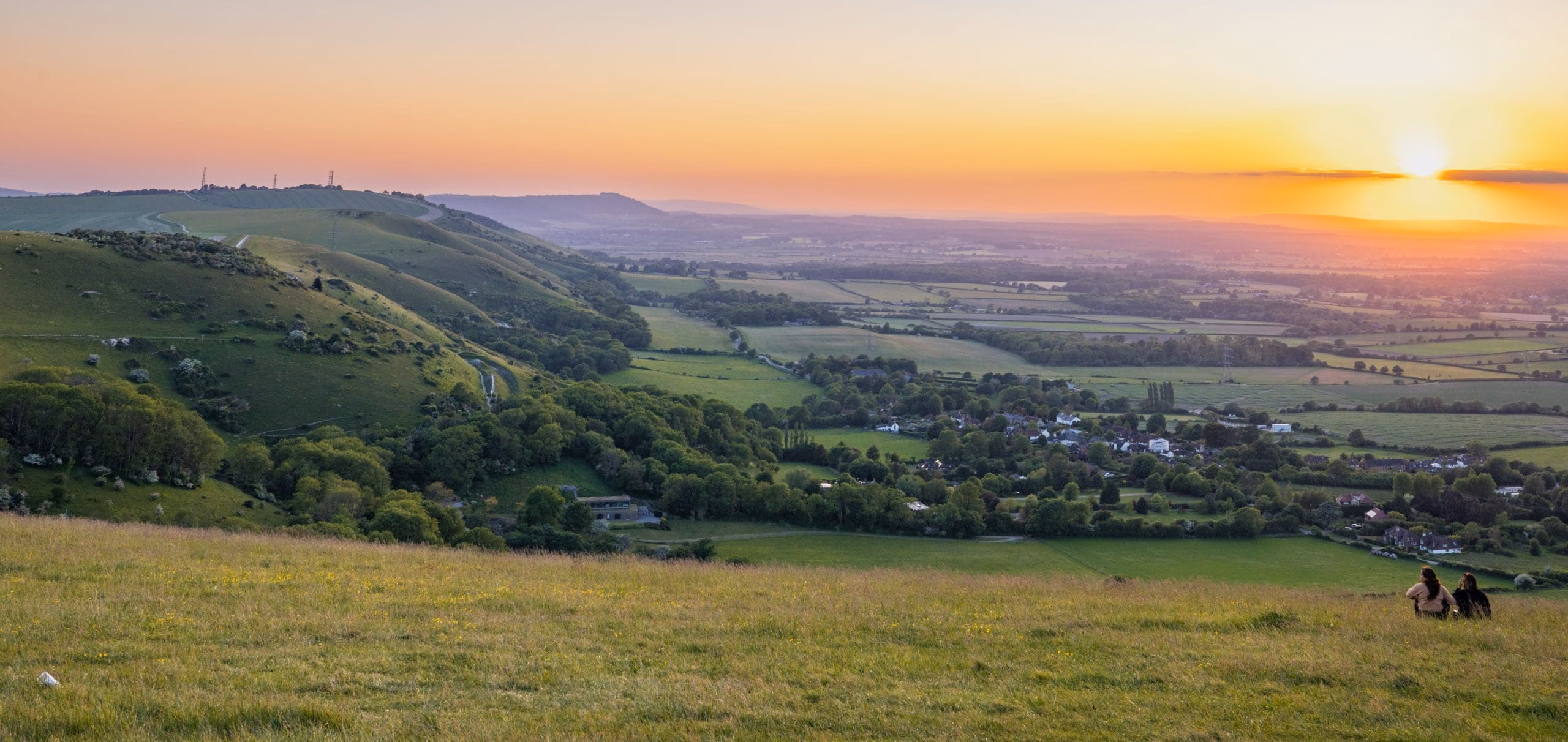A serene landscape at sunset with rolling green hills and a distant village. Two people sit on a grassy slope, enjoying the warm, orange sky.