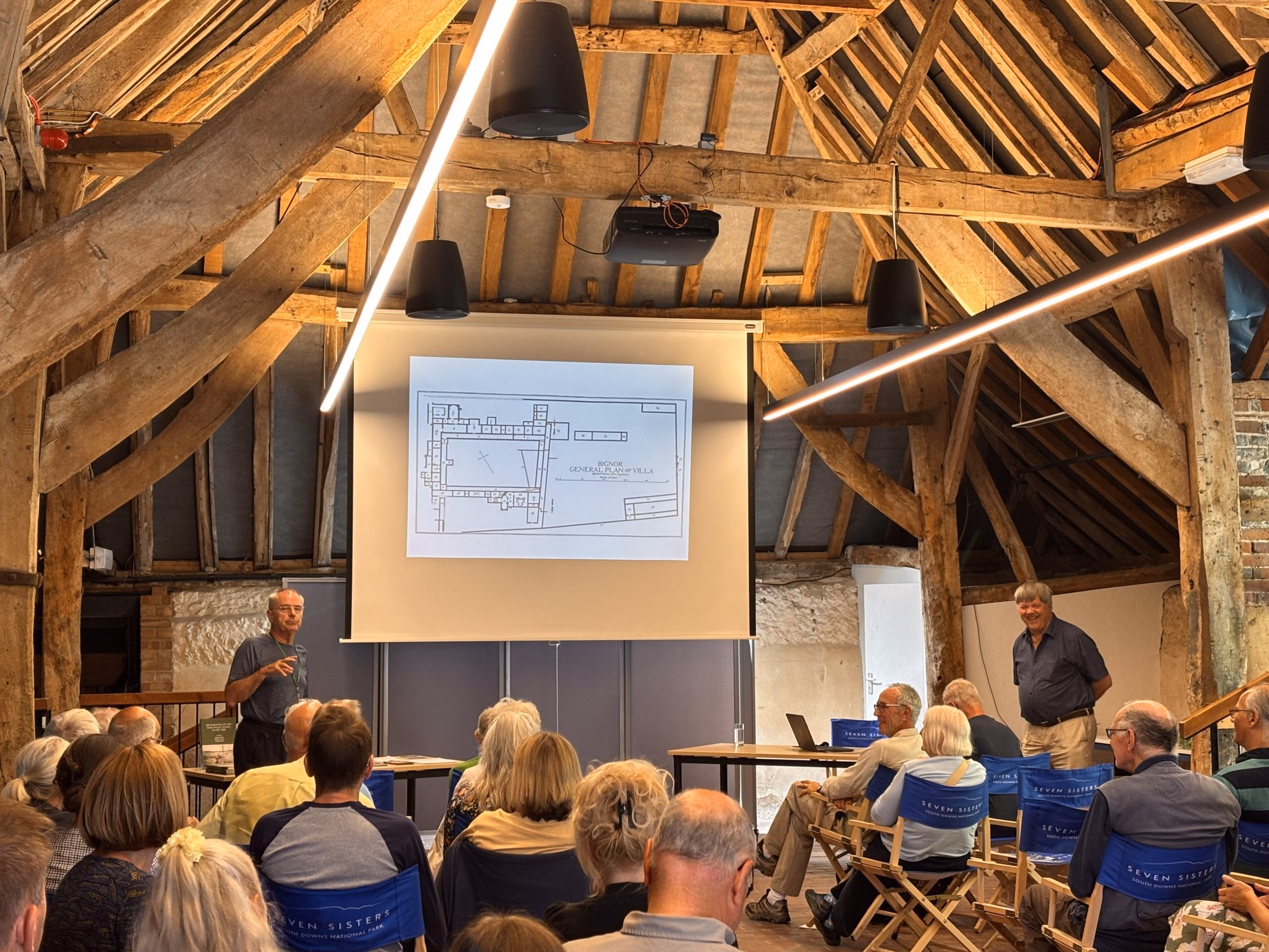 A crowd being addressed by two speakers in a barn.