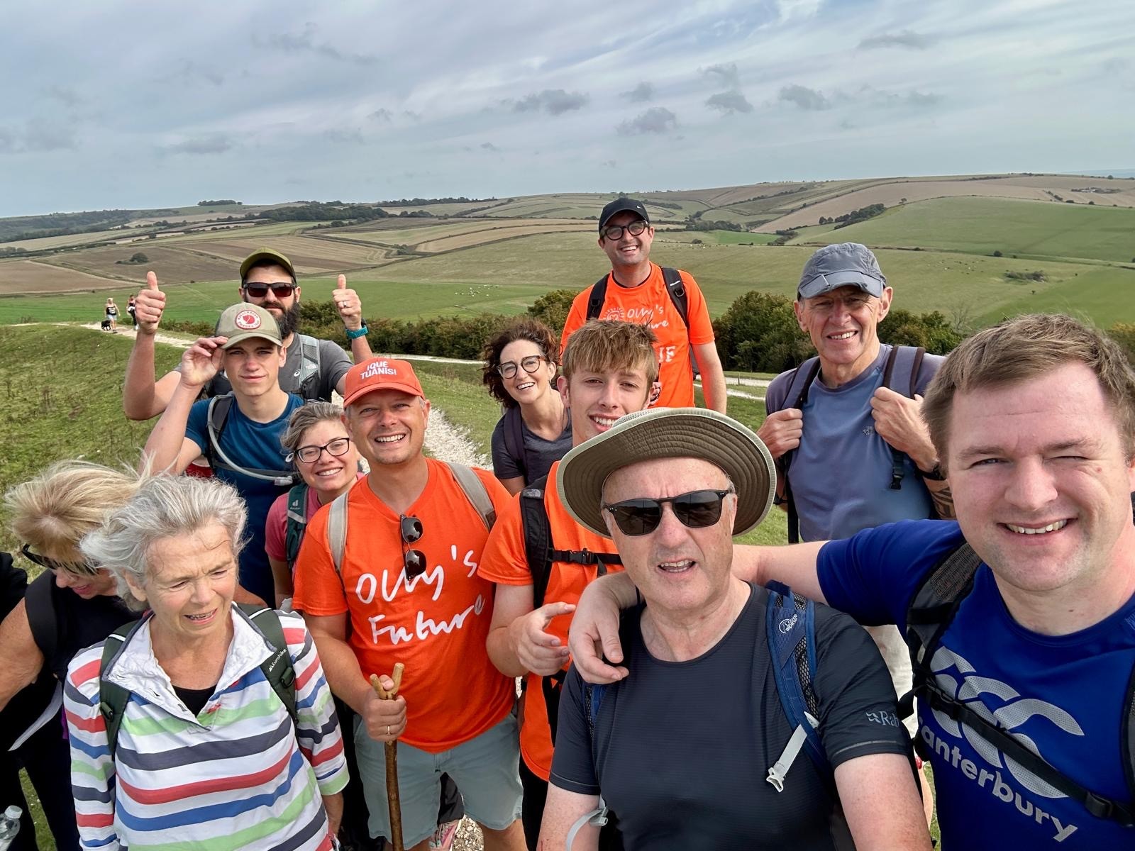 A group of walkers atop a hill on the South Downs smiling for the camera, with some wearing orange t-shirts in support of Oll'ys Future charity.